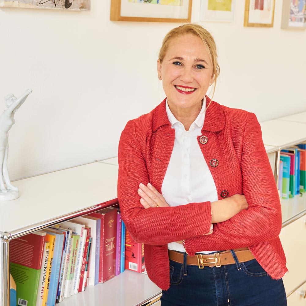 Systemischer Coach Sabine Samonig von SaSa Coaching lehnt in einer roten Lederjacke an einem Bücherregal und lächelt in die Kamera als Symbolbild für ihre "Über mich"-Seite.