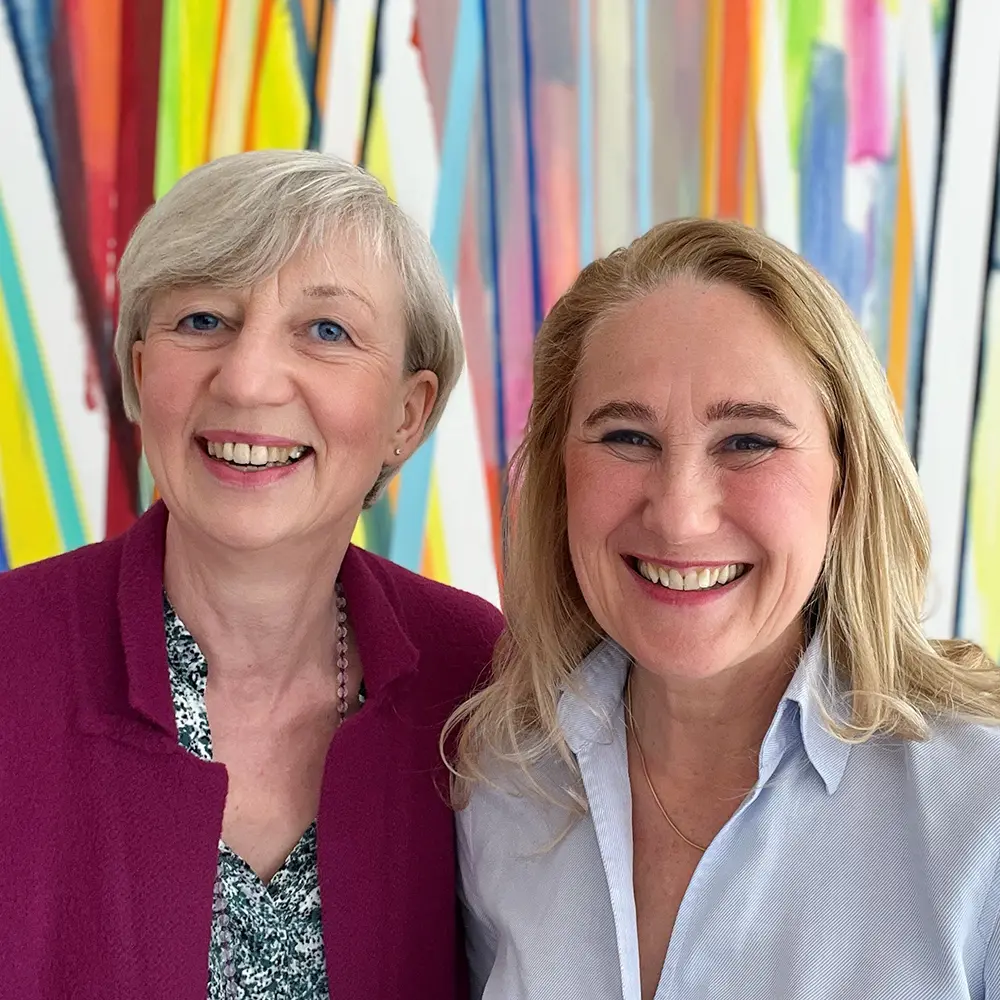 Workshop Trainerinnen Susanne Bosch und Sabine Samonig lächeln vor einem bunten Kunstwerk aus Graffitistreifen in die Kamera.