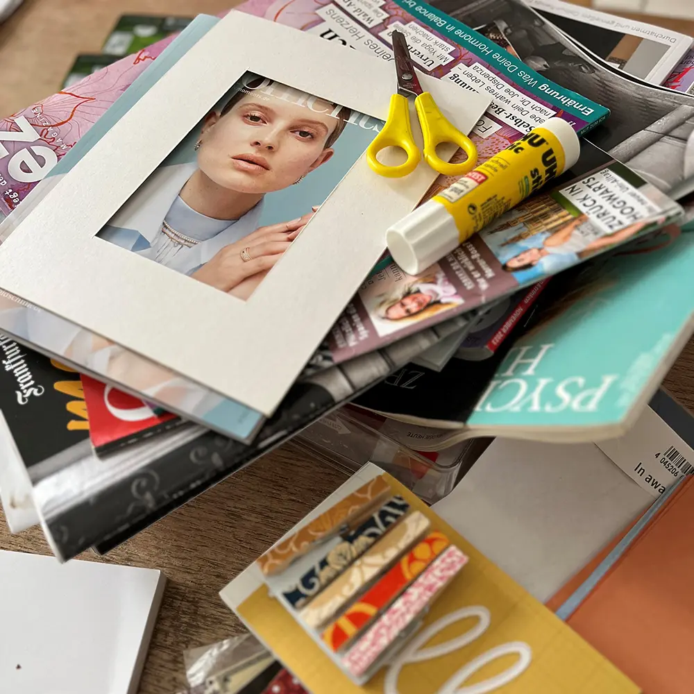 Ein Passpartout, eine gelbe Bastelschere und ein Stift Uhukleber liegen auf einem Stapel Zeitschriften als Symbol für den Workshop "Collagerei – Ideenrausch mit Dame".